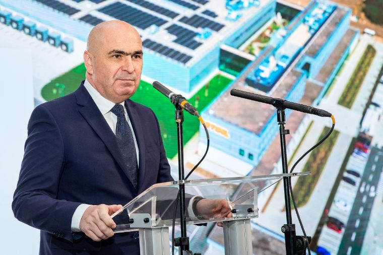 Ilie Bolojan im dunklen Anzug spricht am Rednerpult mit zwei Mikrofonen vor gro&szlig;formatiger Luftaufnahme eines Firmengeb&auml;udes