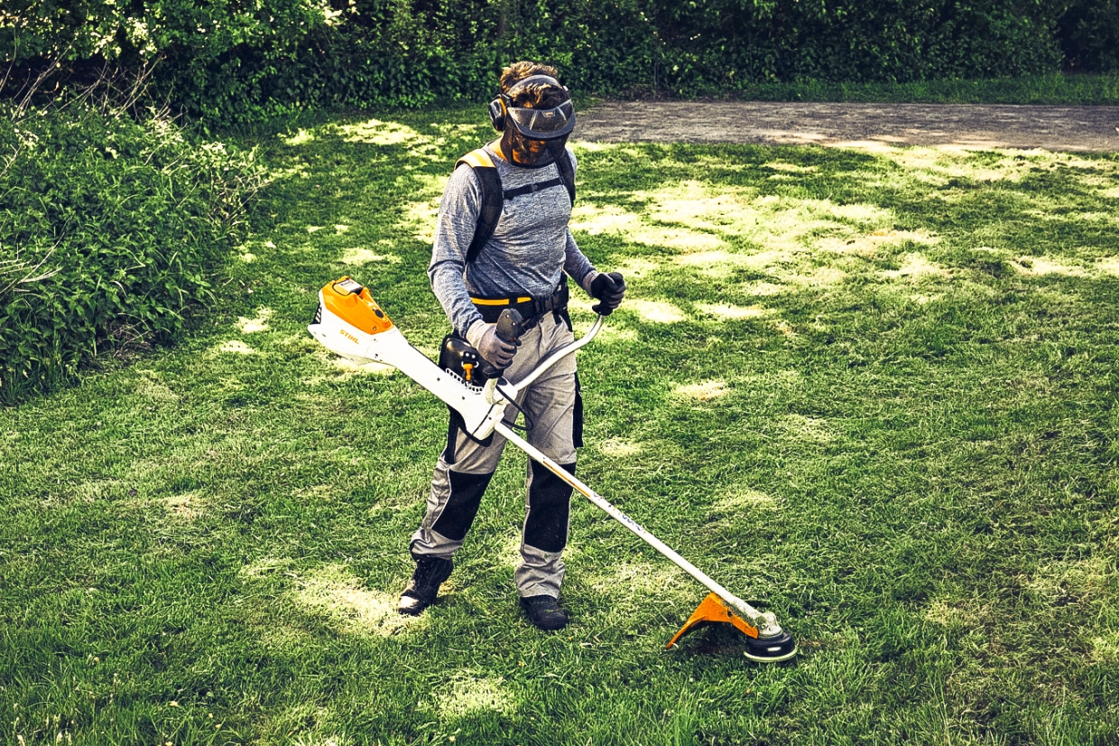 Person mit Schutzhelm, Visier und Gehörschutz arbeitet mit STIHL Freischneider auf sonniger Rasenfläche am Gartenrand