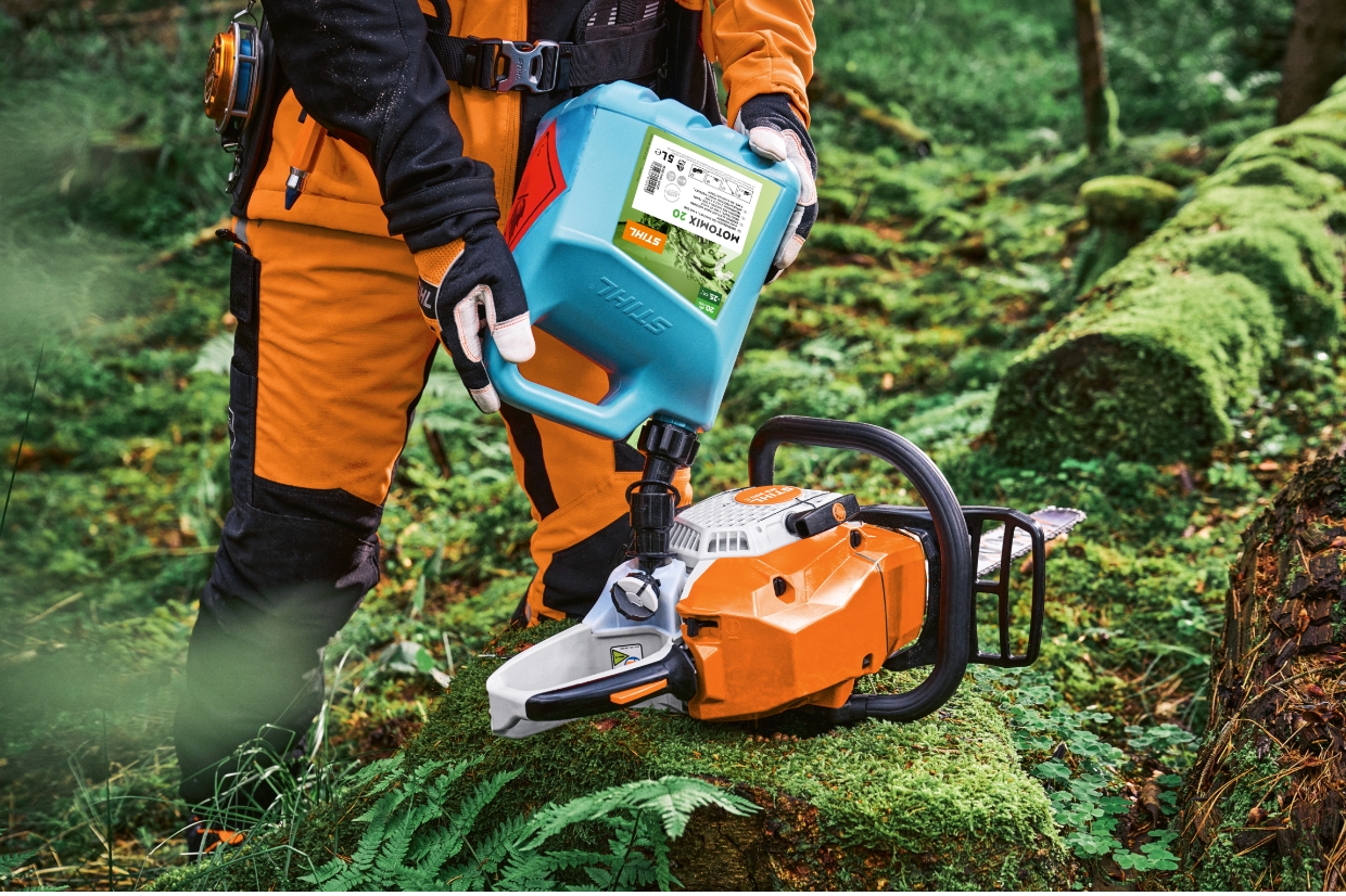 Person in orange Schutzkleidung betankt eine STIHL Motors&auml;ge mit MotoMix Kanister auf moosbewachsenem Baumstamm im Wald.