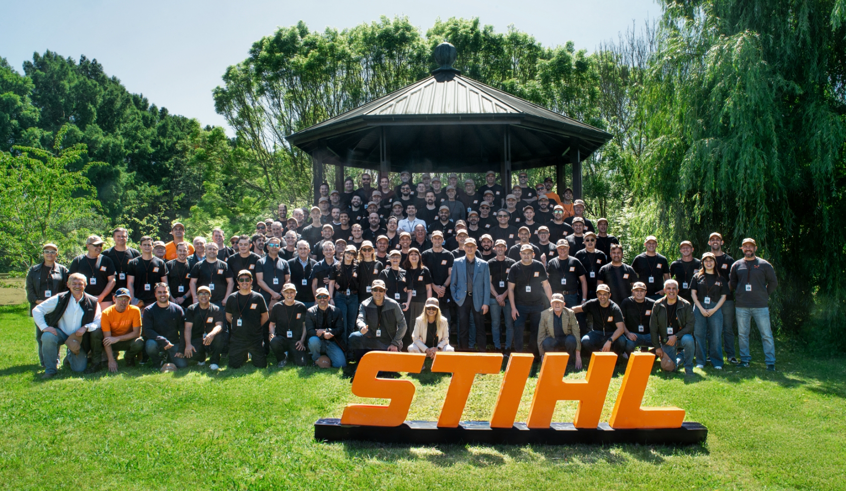 Gro&szlig;e STIHL Belegschaftsgruppe vor Pavillon im Park, alle tragen Firmenkleidung und Ausweise, STIHL Schriftzug im Vordergrund