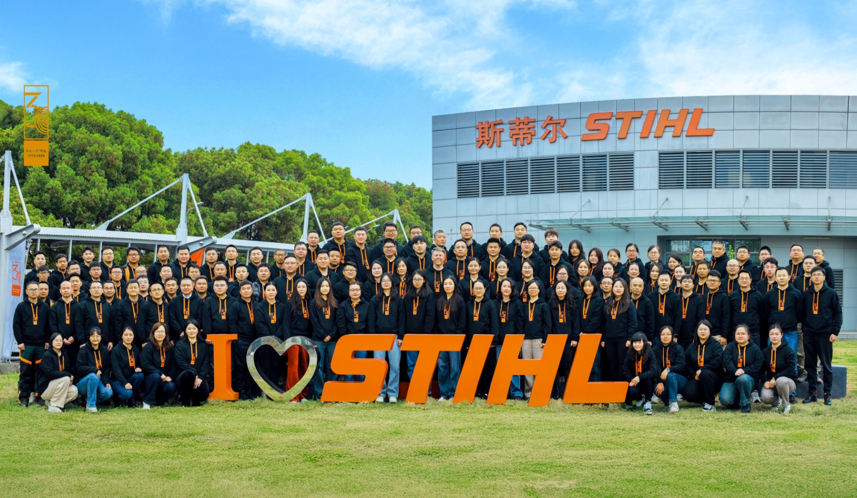Gro&szlig;e STIHL Belegschaft vor Firmengeb&auml;ude mit Schriftzug, alle tragen schwarze Hoodies mit orangenen Kordeln auf gr&uuml;ner Wiese