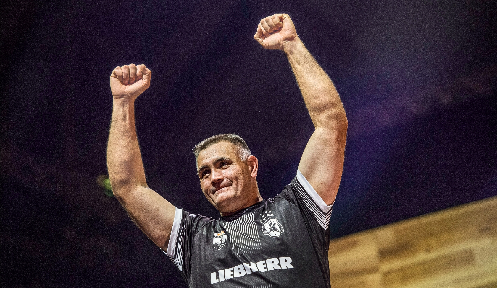 Timbersports Athlet im dunklen Trikot hebt beide Arme auf beleuchteter B&uuml;hne, jubelnde Zuschauer verschwimmen im Hintergrund