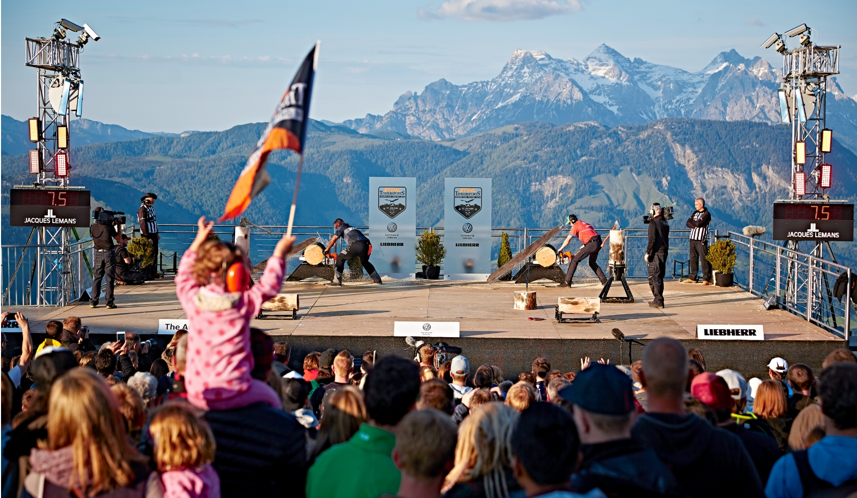 STIHL Timbersports Wettkampf auf Bergb&uuml;hne mit zwei S&auml;geteams, Kind mit Geh&ouml;rschutz und Fahne im Vordergrund, Alpenpanorama