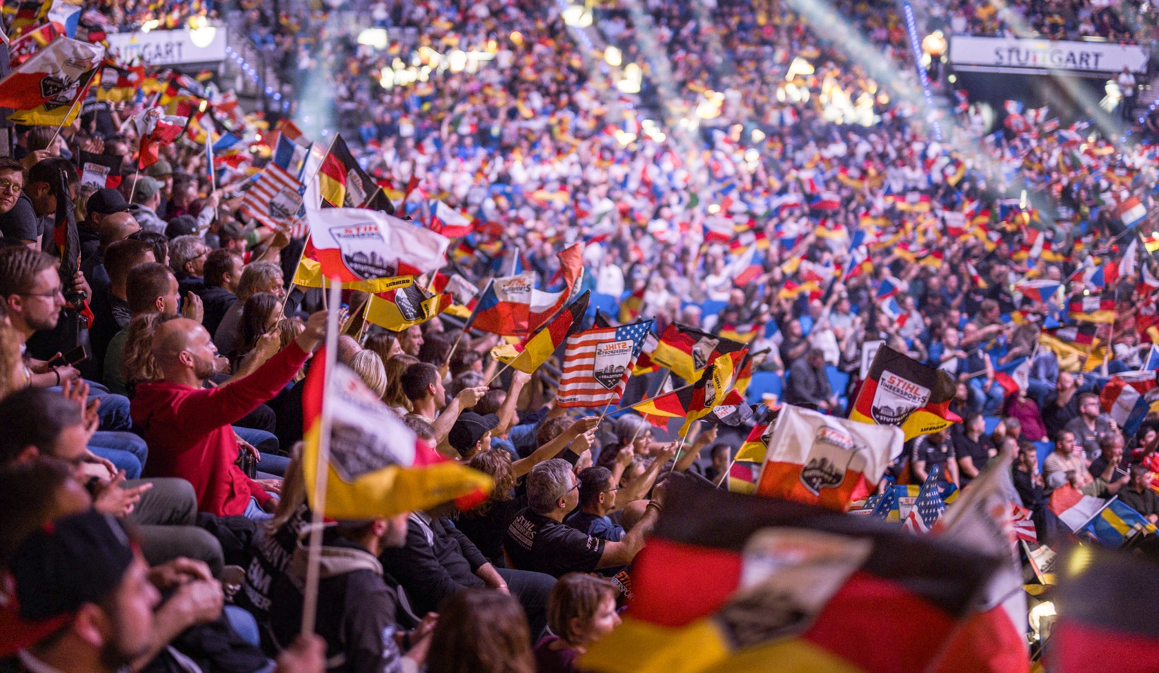 Gro&szlig;e Arena voller STIHL Timbersports Fans, Hunderte schwenken Fahnen verschiedener Nationen, Scheinwerfer beleuchten R&auml;nge