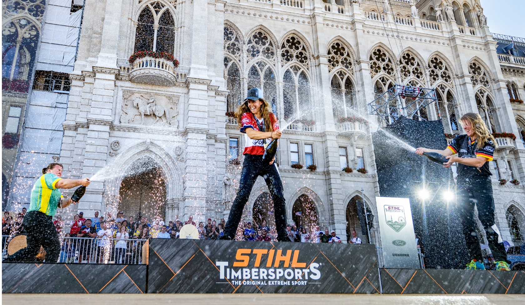 Drei STIHL Timbersports Athletinnen spr&uuml;hen Champagner vor historischer Rathausfassade, Siegerpodest mit Logo im Vordergrund