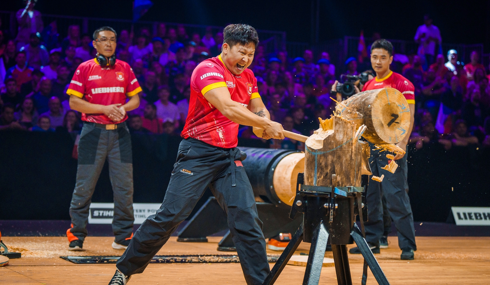 STIHL Timbersports Wettk&auml;mpfer im roten Trikot spaltet mit Axt Holzblock, Teamkollegen und Publikum f&uuml;llen beleuchtete Arena