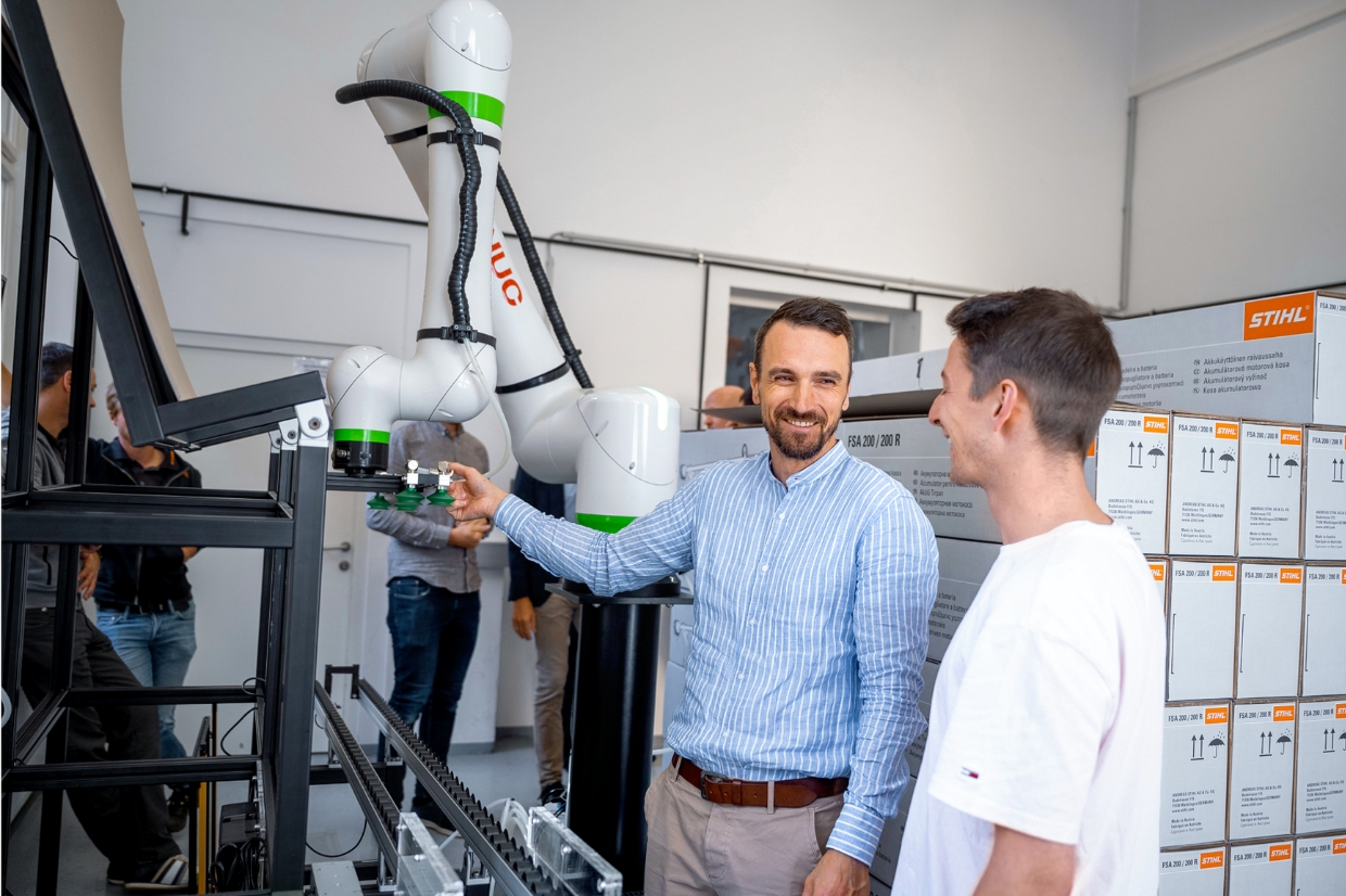 Zwei Mitarbeitende sprechen neben kollaborativem Roboterarm an F&ouml;rderstrecke, Kartons mit STIHL Produkten dahinter gestapelt