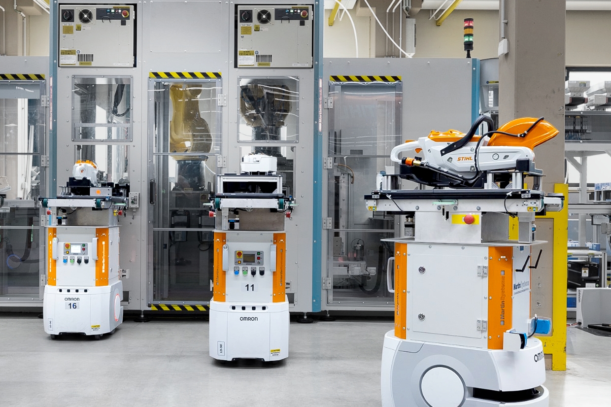 Mehrere autonome Omron Transportroboter mit STIHL Ger&auml;ten fahren vor verglaster Fertigungszelle in moderner Produktionshalle