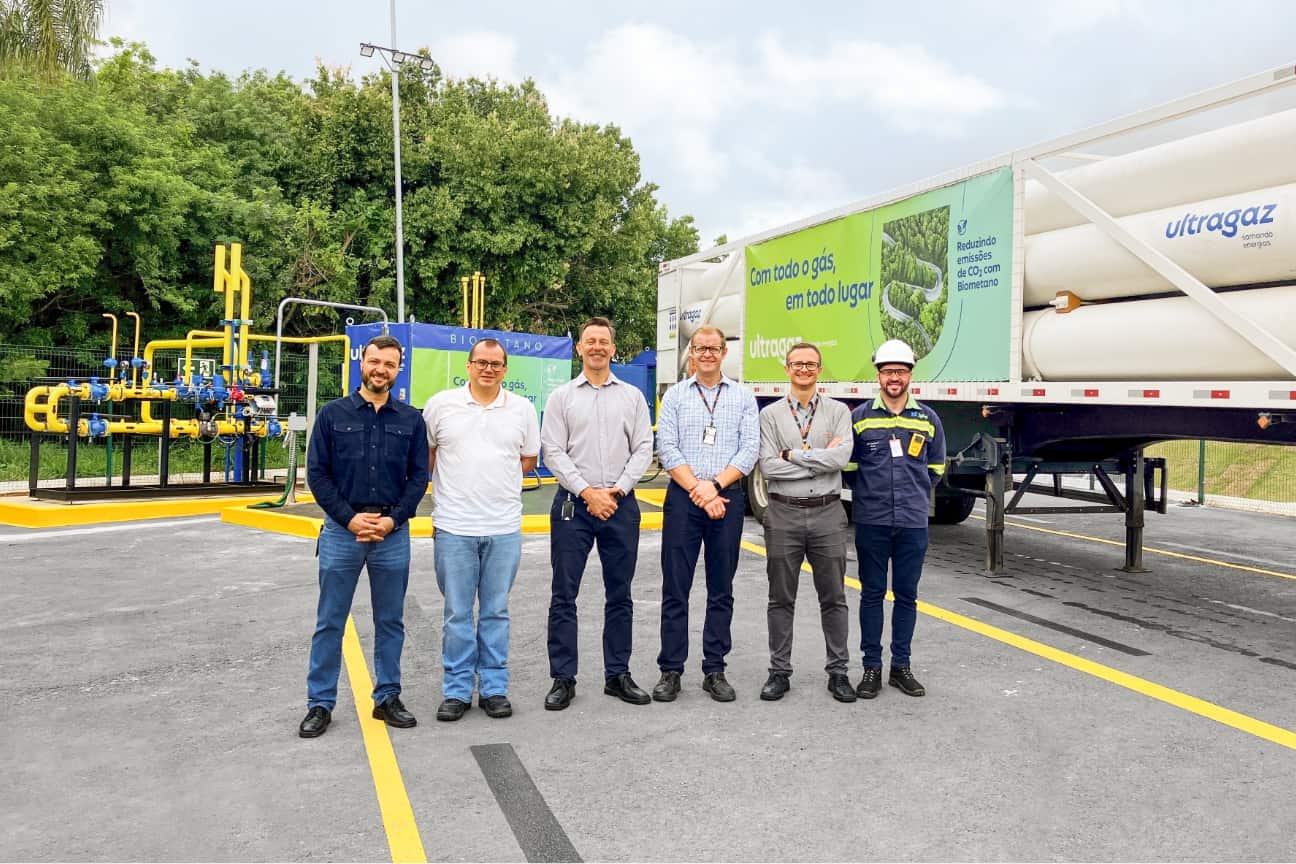 Sechs M&auml;nner stehen vor Biogas Anlage und Ultragaz LKW mit Biomethan Werbung, gelbe Rohrleitungen und B&auml;ume im Hintergrund